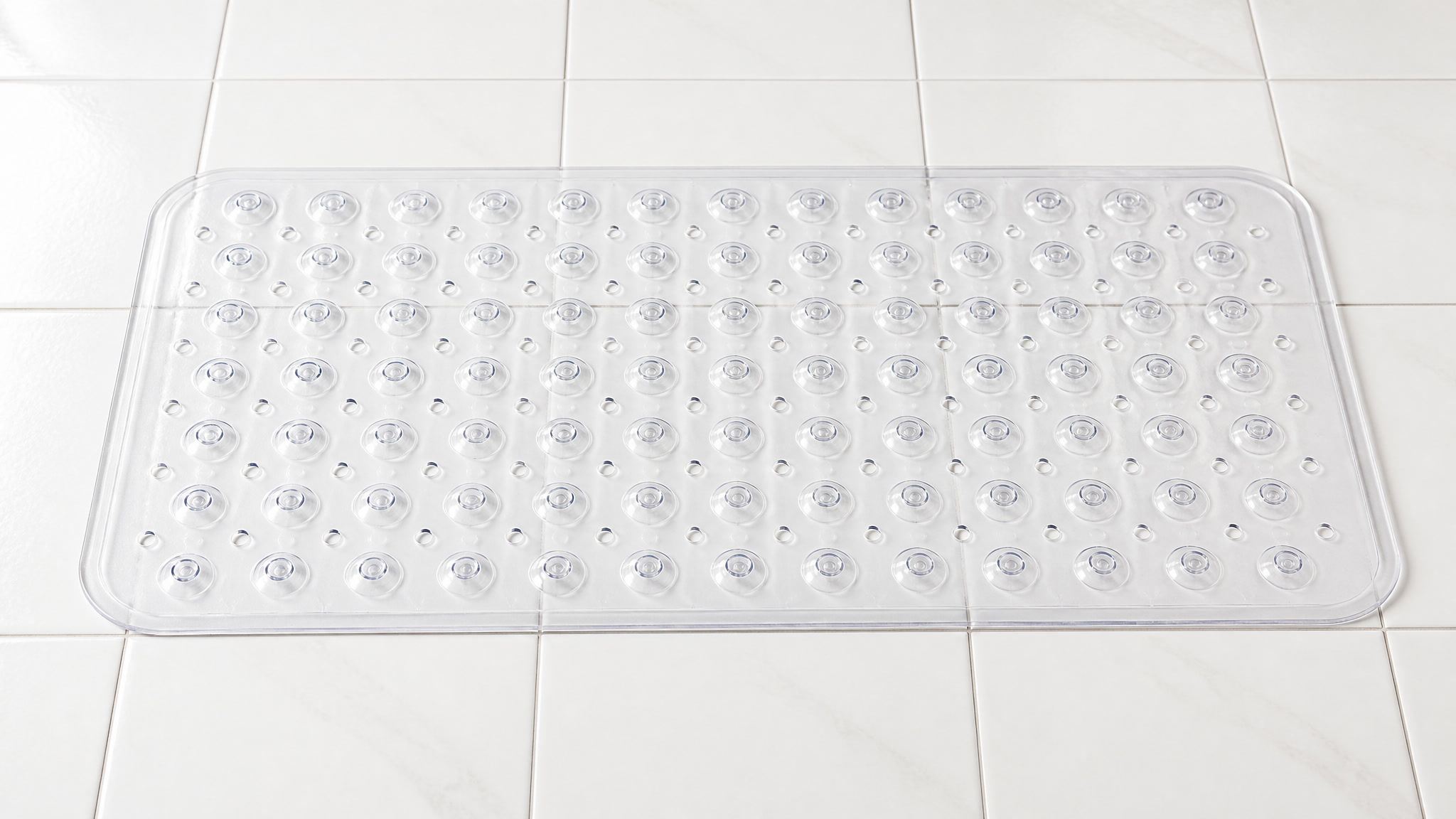 Clear non-slip bathtub mat with suction cups and drain holes laid flat on white tile